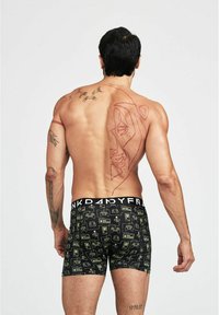 Boxer-briefs noirs avec des motifs de manettes de jeu et d'appareils verts ; taille élastique avec le logo 'PANDY' ; personne avec des tatouages visibles sur le torse.