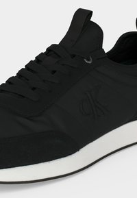 Μαύρο παπούτσι τύπου sneaker χαμηλής κοπής με μαύρα κορδόνια, ανάγλυφο λογότυπο "ck" στο πλάι και λευκή σόλα με μαύρο κάτω στρώμα.