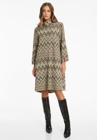 Frau in einem beigefarbenen, zigzag-musterierten knielangen Kleid mit langen Ärmeln und schwarzen knielangen Stiefeln, die vor einem weißen Hintergrund steht.