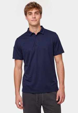 Giovane uomo in piedi che indossa una polo blu navy tinta unita a maniche corte con pantaloni scuri su sfondo bianco.