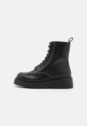 Botines de cuero negro con suela plataforma, diseño con cordones, detalle de costuras y una etiqueta en forma de lazo en el talón para facilitar su colocación.