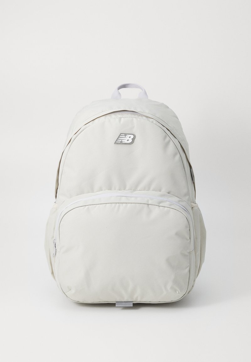 Lys grå ryggsekk med en jevn tekstur, med et håndtak på toppen, en glidelåslomme foran, og en New Balance-logo på forsiden.