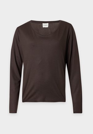 Femme debout, face à l'avant, portant un haut à manches longues marron foncé, un pantalon beige clair et des chaussures à lacets marron foncé sur un fond uni.