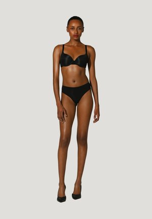 Femme debout devant un fond uni, portant un soutien-gorge noir, des sous-vêtements noirs et des talons hauts noirs, face à l’avant, bras détendus.