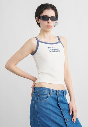 Noor, kes kannab musta kolmnurksete klaasidega päikeseprille, valget tankitoppi sinise ääre ja tekstiga ning kõrge vöökohaga sinist denimist seelikut, millel on nööbid.