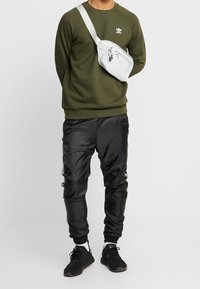 Grå axelremsväska, olivgrön sweatshirt, svarta satäng-joggers och svarta sneakers med vita accenter, stående mot en neutral bakgrund.