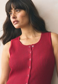 Red ribbed sleeveless top with a round neckline, featuring a front button closure with silver eyelets and a soft texture.