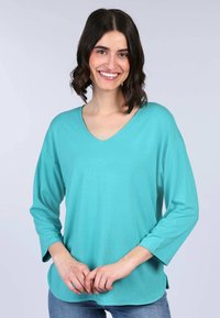 Türkisfarbener V-Ausschnitt-Oberteil mit 3/4-Ärmeln aus weichem Stoff. Es besitzt einen lockeren Schnitt und einen abgerundeten Saum, kombiniert mit hellblauen Jeans.