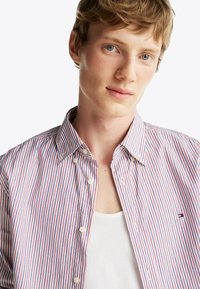 Camisa de rayas en rojo, blanco y azul, con cuello de botón y botones blancos. Llevada sobre una camiseta de tirantes blanca.
