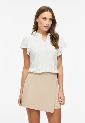 Kobieta ubrana w białą, ażurową bluzkę z krótkim rękawem i falbaniastego kołnierza oraz beżową asymetryczną mini spódnicę, stojąca na białym tle.