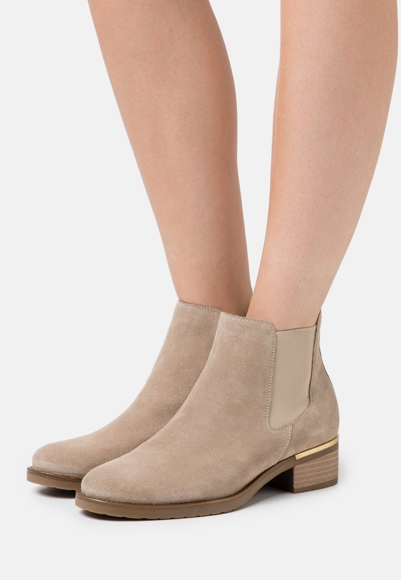 Bottines en daim beige avec panneaux latéraux élastiques et talons bas empilés, portées sur jambes nues sur fond blanc.
