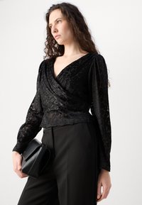 Blusa nera avvolgente con maniche lunghe, motivo testurizzato e scollatura a V profonda. Abbinata a pantaloni neri e una piccola borsa nera.