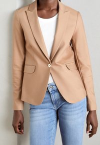 Blazer beige en tissu lisse, avec un seul bouton doré, deux poches avant et des revers crantés, porté sur un haut blanc côtelé.