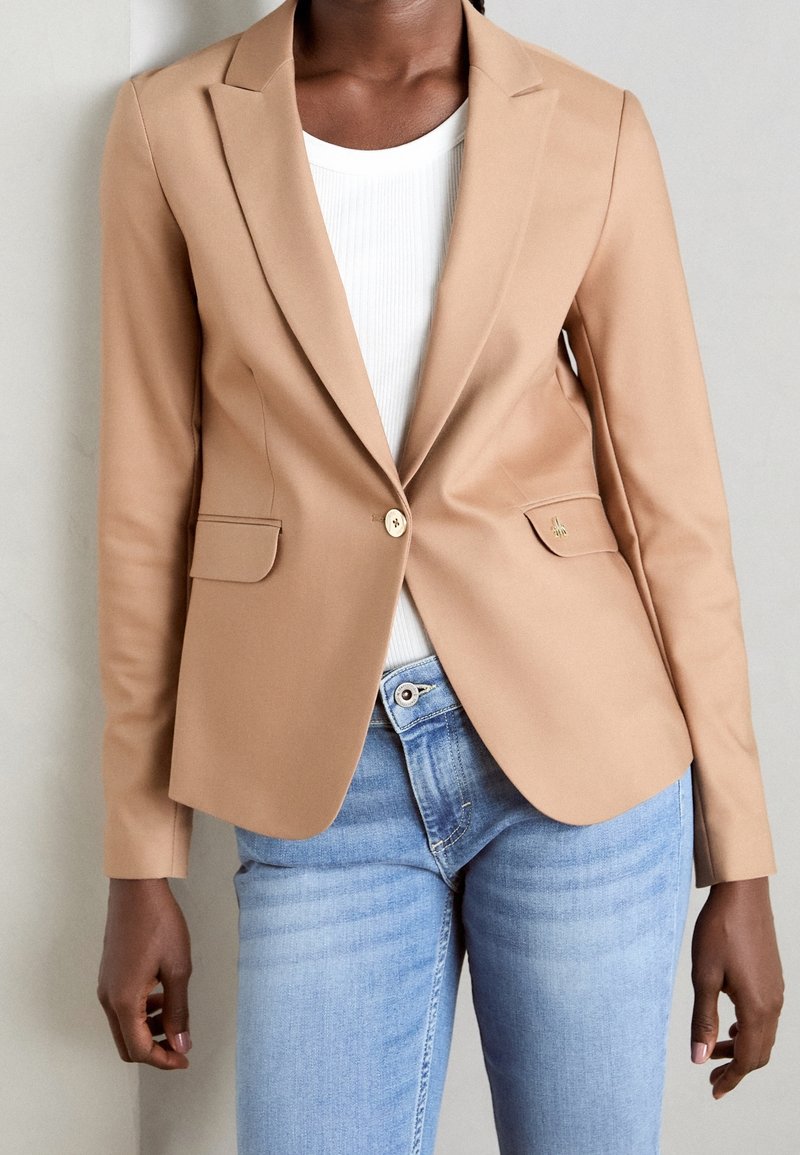 Blazer beige en tissu lisse, avec un seul bouton doré, deux poches avant et des revers crantés, porté sur un haut blanc côtelé.