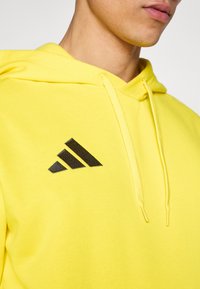 Κίτρινο hoodie από βαμβάκι με υφασμάτινη υφή. Διαθέτει μαύρο λογότυπο με τρεις ρίγες μπροστά και κουκούλα με κορδόνι.