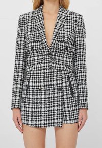 Kvinna som bär en svart- och vitrutad kort blazer och matchande högmidjad kjol med knapp- och fickdetaljer.