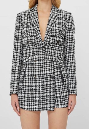 Femme portant un blazer court à carreaux noir et blanc et une jupe taille haute assortie avec des détails de boutons et de poches.