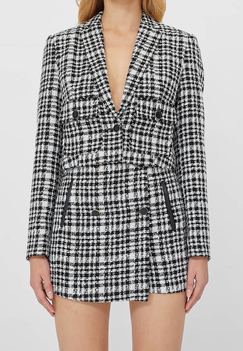 Kvinna som bär en svart- och vitrutad kort blazer och matchande högmidjad kjol med knapp- och fickdetaljer.