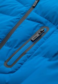 Sininen toppatakki, jossa tikattu pinta. Varustettu mustalla vetoketjulla ja erottuvalla sinisellä vetoketjunauhalla vinossa kohdassa.