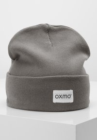 Siva pletiva kapa s mekanim teksturom, koja ima preklopeni rub i bijelu oznaku s natpisom "oxmo" ispisanim u crnoj boji.