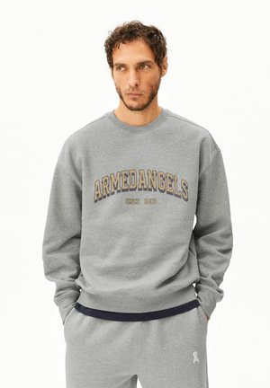 Mann mit lockigem Haar trägt grauen Pullover mit dem Text "ARMEDANGELS since 2007" und passende graue Jogginghose, Hände in den Taschen.