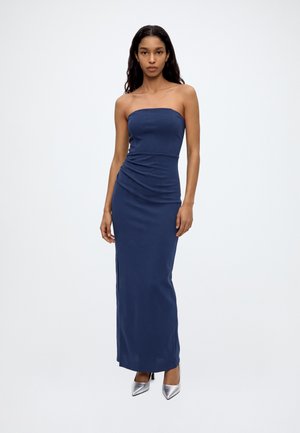Un mannequin se tient devant un fond blanc uni, vêtu d’une robe bustier bleu marine longue jusqu’au sol et de chaussures à talons argentées à bout pointu.