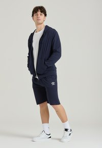 Felpa blu navy con zip abbinata a pantaloni corti blu navy con logo bianco, completata da sneakers bianche e calze a fantasia. Design semplice e sportivo.