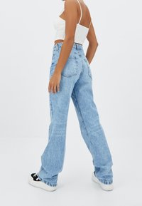 Jeans de mezclilla de pernera ancha en azul claro, con una cintura alta, diseño de cinco bolsillos y dobladillos sin rematar. Combinados con zapatillas en blanco y negro.