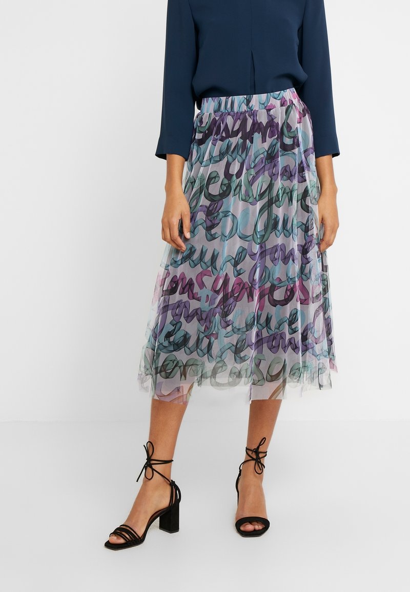 Femme portant une jupe midi en tulle avec un imprimé abstrait multicolore en forme de script, un chemisier bleu marine à manches trois-quarts, et des talons blocs noirs avec lanières à la cheville.