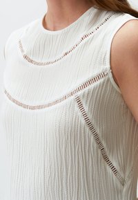 Top senza maniche bianco con texture, caratterizzato da ritagli orizzontali e dettagli di cucitura intricati lungo il colletto e i lati.
