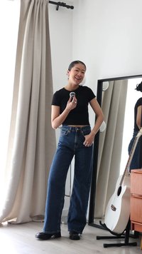 Jonge vrouw in een zwart crop top en wijd uitlopende spijkerbroek glimlachend, houdt een make-up kwastje vast bij een vloerspiegel met een witte gitaar in de buurt.
