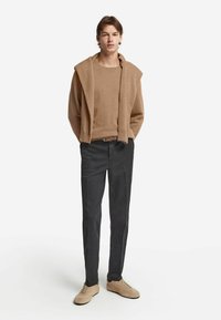 Beige Strickpullover mit angegliedertem Kapuzen, der über den Schultern fällt, kombiniert mit dunkelgrauen Hosen und hellbeigen Wildlederschuhen. Schlichtes Design, lässige Passform.