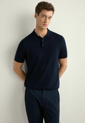 Młody mężczyzna w granatowym polo z krótkim rękawem i spodniach stoi na tle jasnej, gładkiej ściany z rękami za plecami.