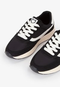 Deportivas negras con un diseño elegante, materiales de gamuza y tela, cordones blancos y una plantilla crema. Acentuadas con un detalle de raya blanca.