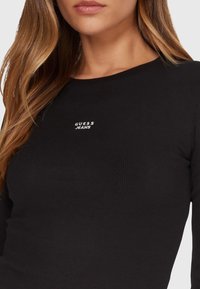 Top nero a maniche lunghe a coste con scollo rotondo, con logo bianco "GUESS JEANS" al centro del petto. Texture morbida.