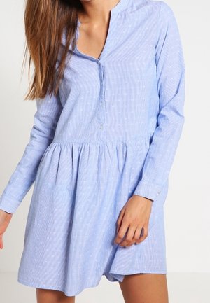 Femme portant une robe bleue claire à manches longues, boutonnée, avec des motifs discrets de points blancs et un détail de taille froncée.