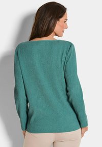 Eine Frau trägt ein strukturiertes, türkisfarbenes Langarmoberteil und beige Hosen. Sie steht mit dem Rücken zur Kamera und schaut nach rechts vor einem weißen Hintergrund.