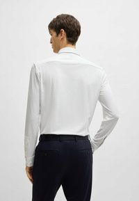 Camicia bianca a maniche lunghe con una finitura testurizzata, dotata di colletto e polsini classici. Abbinata a pantaloni blu scuro, vista da dietro.