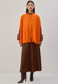 Cape orange en tricot avec fermeture à boutons sur le devant et fentes latérales, associée à une longue jupe marron et des bottes marron à talons montant jusqu'aux genoux, textures lisses.