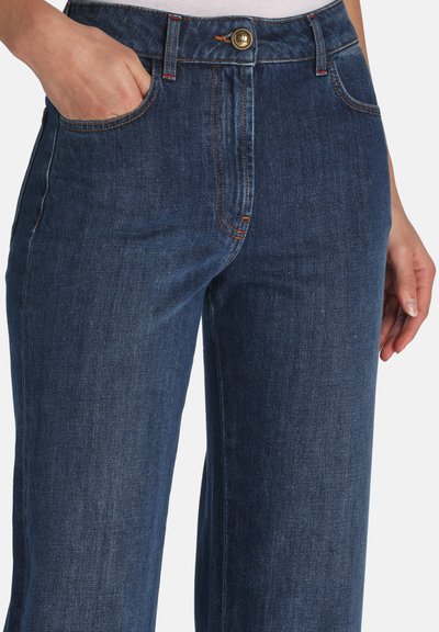 Tamnoplave traperice ravnog kroja s kopčanjem na tipku od mesinga, crvenim šavovima za naglasak i prednjim džepovima.