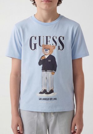 T-shirt azzurro chiaro con la scritta "GUESS" e un orso che indossa una giacca nera, pantaloni a righe e scarpe da ginnastica, in piedi al centro.