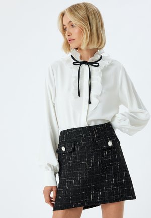 Witte blouse met gerimpelde kraag en zwarte strik, gecombineerd met een zwarte tweed minirok met voorkruiszakken en witte knoopaccenten.