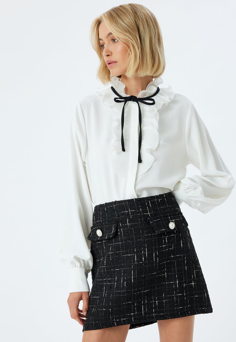 Camicia bianca con colletto arricciato e cravatta nera, abbinata a una mini gonna nera in tweed con tasche frontali e dettagli con bottoni bianchi.