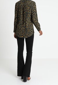 Das Hemd hat eine olivgrüne Basis mit einem Leopardenmuster in Schwarz und Orange, langen Ärmeln und einer lässigen Passform. Dazu werden schwarze Schlaghosen getragen.
