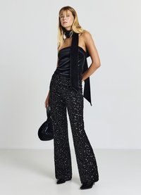 Schwarzes schulterfreies Satin-Top mit langem Nackenschal, kombiniert mit ausgestellten schwarzen Paillettenhosen. Das Model hält eine kleine schwarze Tasche und trägt Absätze.