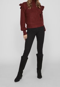 Frau in einem burgunderfarbenen Strickpullover mit Rüschenschultern, schwarzen Skinny-Hosen und schwarzen kniehohen Stiefeln, die vor einem schlichten Hintergrund steht.