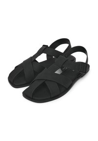 Schwarze Sandalen aus flexiblem Material, mit überkreuzten Riemen, einer verstellbaren Schnalle, offenem Zehenbereich und flacher Sohle.