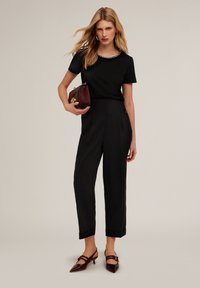 Zwarte top met korte mouwen en een kralen halslijn, gecombineerd met zwarte high-waisted broeken. Het model houdt een bordeauxrode handtas vast en draagt bruine hakken.