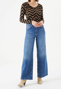 Brown and black zigzag patterned long-sleeve top paired with high-waisted light blue wide-leg jeans and beige ankle boots.