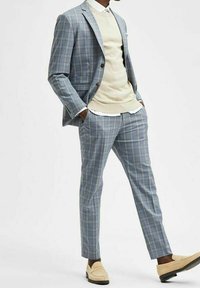 Herrjacka i rutigt mönster i ljusblått med beige accenter; skräddarsydd passform på jackan och slim fit byxor; kombinerad med en beige tröja och loafers.